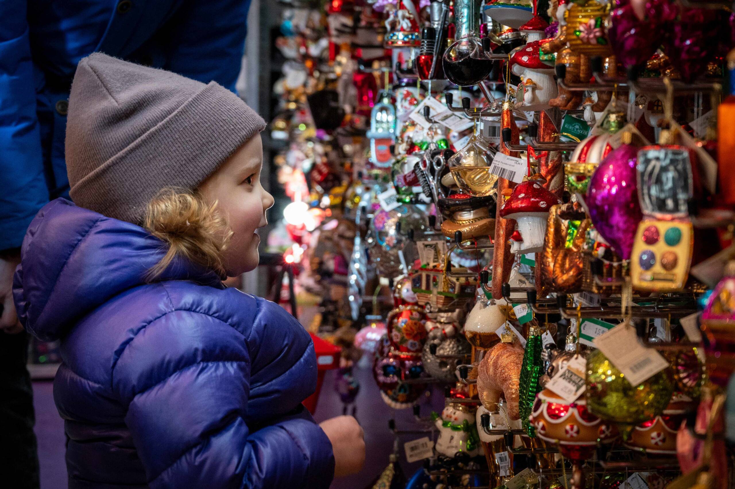 Een jong kind in een paarse winterjas en grijze muts kijkt vol verwondering naar een kleurrijk assortiment van kerstornamenten, uitgestald in een winkel. De ornamenten zijn speels en gevarieerd, met verschillende vormen zoals paddenstoelen en kerstversieringen, wat een feestelijke sfeer creëert.